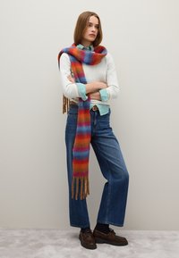 Multikleurig gestreepte sjaal met franjes, gedrapeerd over een lichte trui, gecombineerd met blauwe high-waisted jeans en bruine veterschoenen.