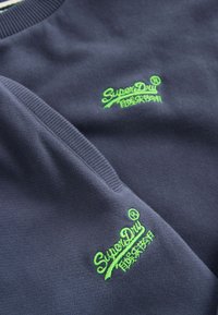 Tela azul oscuro con logotipos "SuperDry" bordados en verde brillante y caracteres japoneses, mostrando una sudadera y una manga dobladas.