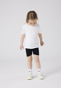 T-shirt en coton blanc associé à un short noir, arborant un design simple. L'enfant porte des baskets blanches et des chaussettes avec un logo noir.