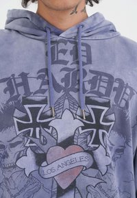 Hombre con una sudadera morada con cruces negras, una pancarta roja con el texto "LOS ÁNGELES" y diseños de calaveras en la tela.