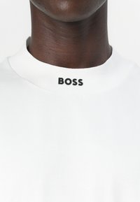 Hvit bomulls t-skjorte med ribbet rund hals og svart "BOSS"-logo ved kragen. Glatt tekstur, minimalistisk design.