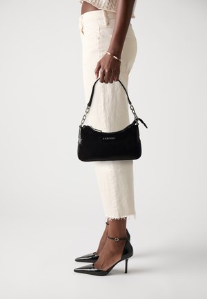 Femme portant un jean cropped crème, des escarpins noirs à bout pointu, tenant un sac à main en cuir verni noir avec une chaîne.