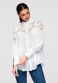 Femme portant un chemisier blanc boutonné avec des détails en dentelle délicate sur les épaules et des manches bouffantes, associé à un jean bleu.