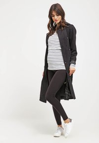 Femme enceinte portant un long manteau noir, un haut blanc rayé, des leggings noirs et des baskets blanches, debout devant un fond uni.
