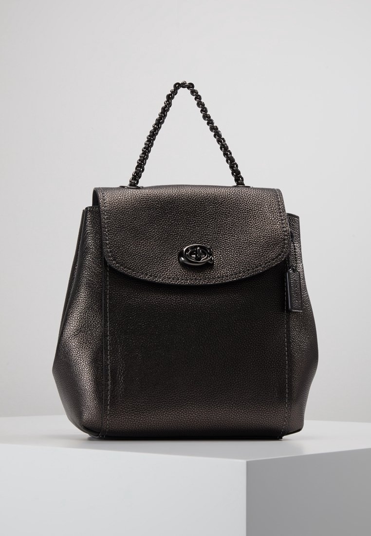 Sac à main en cuir noir texturé avec rabat et fermeture à verrou tournant, poignée courte en chaîne, exposé sur un piédestal blanc sur fond gris.