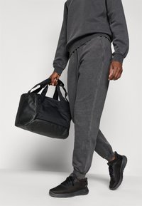 Črna torba za športno opremo z dvema ročajema, gladka tekstura, kontrastni detajli iz tkanine; oseba nosi temno sivo trenirko in superge.