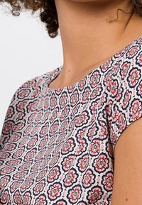 Kortærmet bluse med et blomstermotiv i rød og marineblå på en cremefarvet baggrund, med en glat stofstruktur og rund halsudskæring.