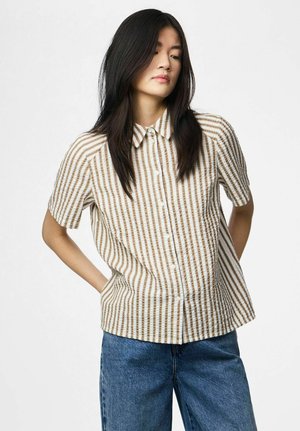 Jonge vrouw met lang zwart haar, gekleed in een beige-wit gestreept overhemd met korte mouwen en een knoopsluiting, en een blauwe spijkerbroek, met de handen achter haar rug.