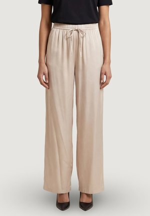 Pantaloni beige a gamba larga con vita elasticizzata e coulisse, abbinati a top nero e tacchi neri con punta a stiletto, in piedi su sfondo chiaro.
