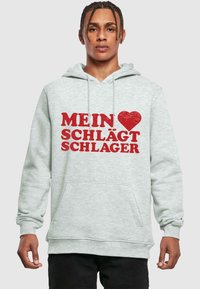 Ljusgrå hoodie med framficka, med fet röd text som läser "MEIN ♥ SCHLÄGT SCHLAGER." Designad med dragsko i huvan och ribbade mudd. 