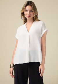 Blouse blanche ample à manches courtes retroussées, design en V, fabriquée en tissu léger, associée à un pantalon noir.