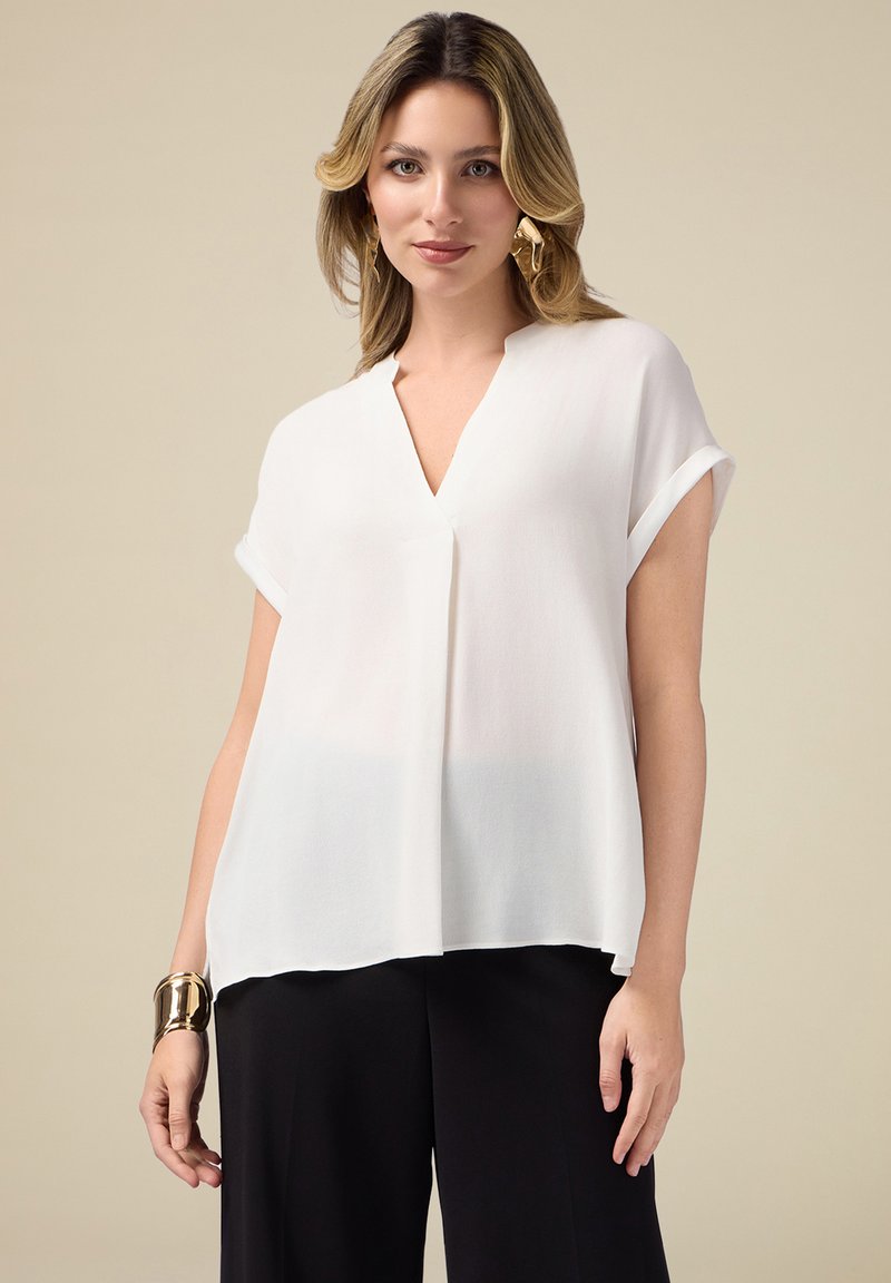 Blouse blanche ample à manches courtes retroussées, design en V, fabriquée en tissu léger, associée à un pantalon noir.