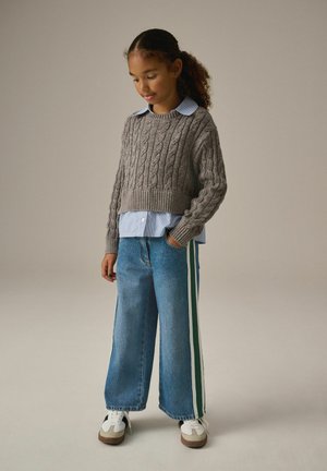 Grå kabelstickad kort tröja över en ljusblå skjorta med krage, kombinerad med vida blå jeans med gröna sidostriper och vita sneakers.