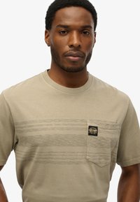 Maglietta beige a maniche corte in cotone, con un motivo a righe texturizzato e una tasca frontale con etichetta sul lato sinistro.