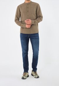 Camicia marrone a maniche lunghe con colletto rotondo e tre bottoni; tasca anteriore. Abbinata a jeans blu scuro e scarpe sportive beige a fantasia.