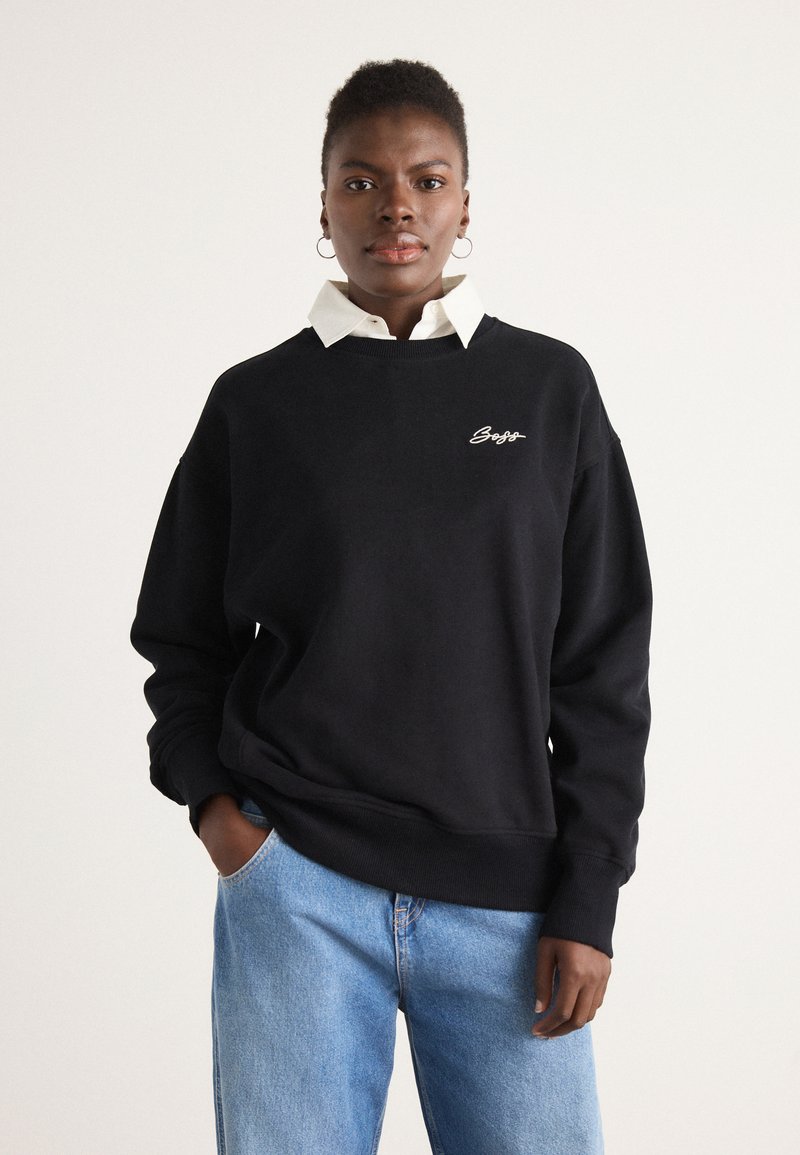 Sudadera de algodón negra con un corte relajado, que presenta un logo bordado en blanco en el pecho, combinada con una camisa blanca con cuello debajo.