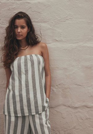 Femme aux longs cheveux bouclés portant un haut bustier à rayures verticales gris et blanc et un pantalon assorti, debout contre un mur beige texturé.