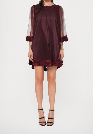 Kobieta ubrana w przezroczystą bordową sukienkę z kwiatową ozdobą na rękawach i dole, w połączeniu z czarnymi sandałami na wysokim obcasie z paskami.