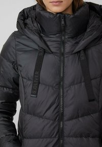 Czarna pikowana kurtka z wysokim kołnierzem, zamek błyskawiczny i regulowane sznurki. Posiada przeszywane panele i akcenty logo na paskach.