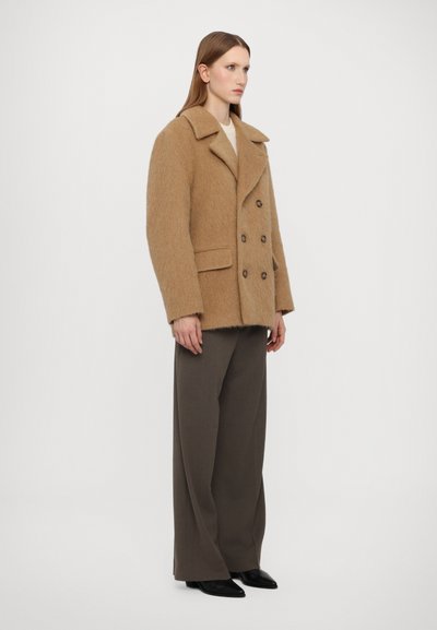 Brązowy płaszcz z wełny z podwójnym zapięciem, dużymi klapami, dwiema kieszeniami z przodu i drewnianymi guzikami, noszony na ciemnych szerokich spodniach.