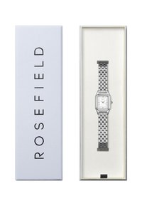 Srebrna pravokutna ručna sat s mrežnom narukvicom, predstavljena u mekoj plavoj kutiji s crnim natpisima. Bijeli brojčanik s minimalističkim oznakama.