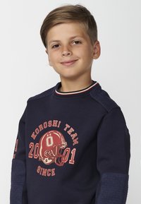 Sudadera de marino con mangas cortas, que presenta un gráfico de fútbol rojo y el texto "KOROSHI TEAM". Cuello acanalado con acentos rojos y blancos, y mangas texturizadas.