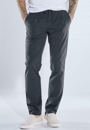 Mann trägt dunkelgraue Hose mit Kordelzug, weiße Slip-On-Schuhe und ein weißes Hemd, die tätowierten Arme sichtbar, steht mit Händen in den Taschen.