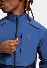 Blue sports jacket made of lightweight, smooth fabric. Features a high collar, zip pocket, and black accents on the shoulders.