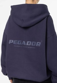 Námo�řnická modrá mikina s kapucí vyrobená z měkké látky, na zadní straně s velkým matným logem "PEGADOR", prostornou kapucí a uvolněným střihem.