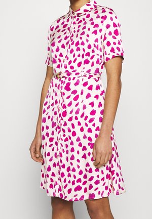Femme portant une robe blanche à manches courtes boutonnée avec des motifs de cœurs et de points roses, accompagnée d'une ceinture assortie à la taille.