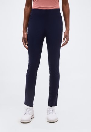 Oseba v mornarsko modrih ozkih hlačah, belih vezalkah in svetlo roza zgornjem brez rokavov na enobarvni podlagi.