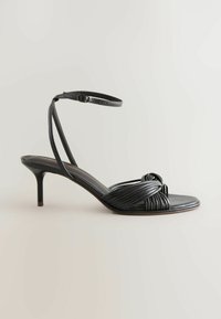Sandale à talon haut en cuir noir avec bride à la cheville et détail noué à l'avant, présentée en profil sur un fond uni.