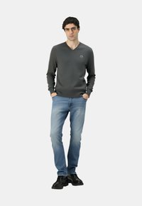 Maglione grigio scuro a maglia con scollo a V, con un piccolo logo sul petto, abbinato a jeans azzurri e calzature nere.