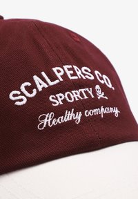 Gorra burdeos con texto bordado en blanco que dice "SCALPEAS CO. SPORTY Compañía saludable". Presenta una forma estructurada y una tela texturizada.