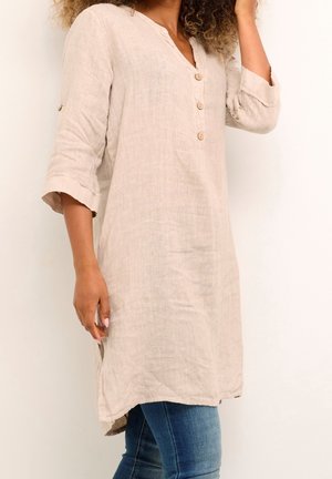 Kobieta ubrana w beżową lnianą tunikę z trzema guzikami i podwiniętymi rękawami, w połączeniu z niebieskimi dżinsami na jednolitym tle.