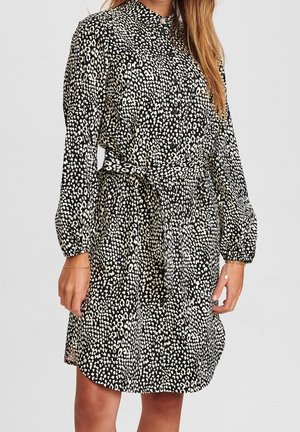 Kobieta ubrana w koszulową sukienkę z długim rękawem w czarno-białe cętki, zapinaną na guziki, z materiałowym paskiem związanym w talii.