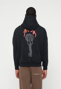 Άνδρας που φορά μαύρη κουκούλα με γραφικό σχέδιο ενός ατόμου που κρατά μια σφαίρα κάτω από κόκκινες φλόγες στην πλάτη, σε συνδυασμό με καφέ παντελόνι με μεγάλες πίσω τσέπες.