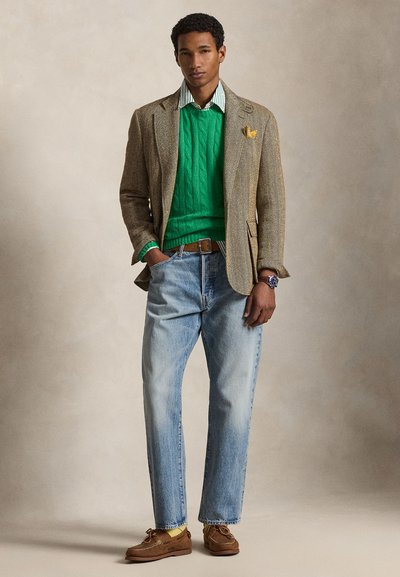 Άνδρας που φοράει πράσινο πλεκτό πουλόβερ με σχέδιο καλώδιο, ριγέ πουκάμισο, καφέ σακάκι σε ύφος ψαροκόκκαλο, ανοιχτό μπλε τζιν και καφέ μοκασίνια, στέκεται με τα χέρια στις τσέπες.