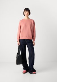 Woman wearing a pink sweatshirt, dark wide-leg jeans, and pink shoes, holding a black handbag against a plain light background.