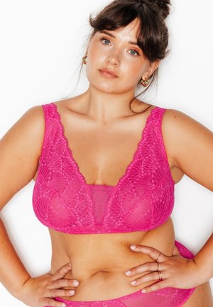 Femme aux cheveux foncés coiffés en chignon, portant des boucles d'oreilles créoles dorées, un soutien-gorge en dentelle rose vif et un bas assorti, posant les mains sur les hanches sur fond blanc.