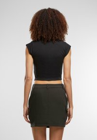 Mujer con cabello rizado que lleva una camiseta sin mangas negra y una falda mini negra con bolsillos laterales, de pie de espaldas sobre un fondo blanco.
