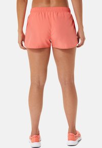 Mujer vestida con pantalones cortos deportivos color coral y zapatillas de deporte a juego, de pie con las piernas y los brazos relajados contra un fondo simple.