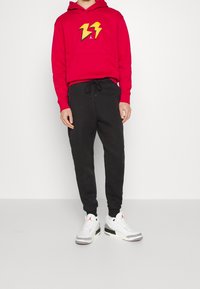 Rode hoodie met geel bliksemlogo, zwarte tapering broek en witte sneakers met zwart en grijze accenten. Zachte katoenmix stof.