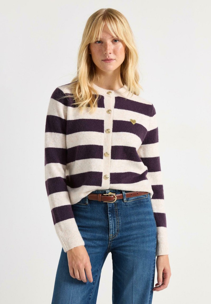 Stribet cardigan i creme og mørk lilla, lavet af blødt stof. Har rund halsudskæring, knaplukning foran og en lille hjerteformet detalje.