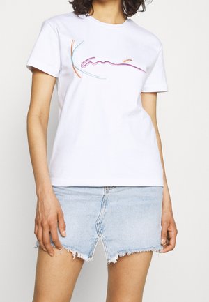 Personne portant un t-shirt blanc avec une broderie abstraite colorée et une mini-jupe en denim bleu clair effilochée.