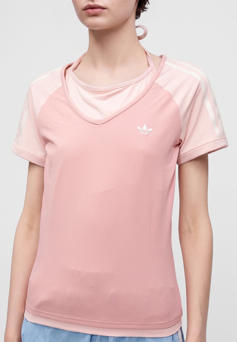 Personne portant un t-shirt rose Adidas à manches courtes avec des bandes blanches sur les manches et un logo sur la poitrine, debout devant un fond uni.