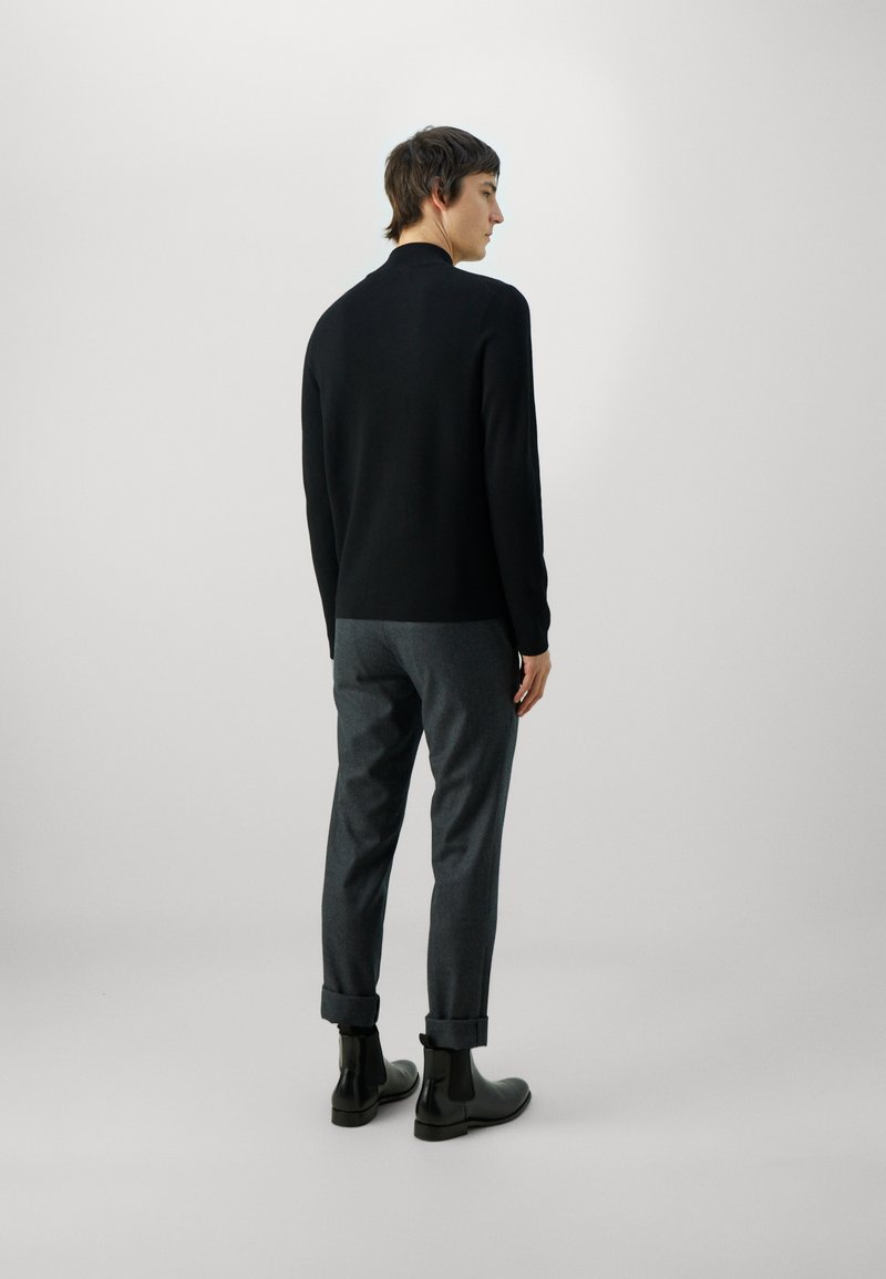 Black knitted sweater with a high neck, paired with grey trousers. Model wears black ankle boots; view from the back. Minimalist design.