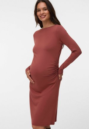 Femme enceinte souriante, portant une robe de maternité ajustée à manches longues couleur rouille avec fronces sur les côtés, debout devant un fond blanc uni.