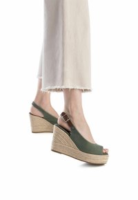 Sandalias verdes de cuña con diseño de punta descubierta, hechas de tela. Presentan un tacón de yute trenzado y una correa ajustable con hebilla. Se ven pantalones con dobladillo deshilachado.