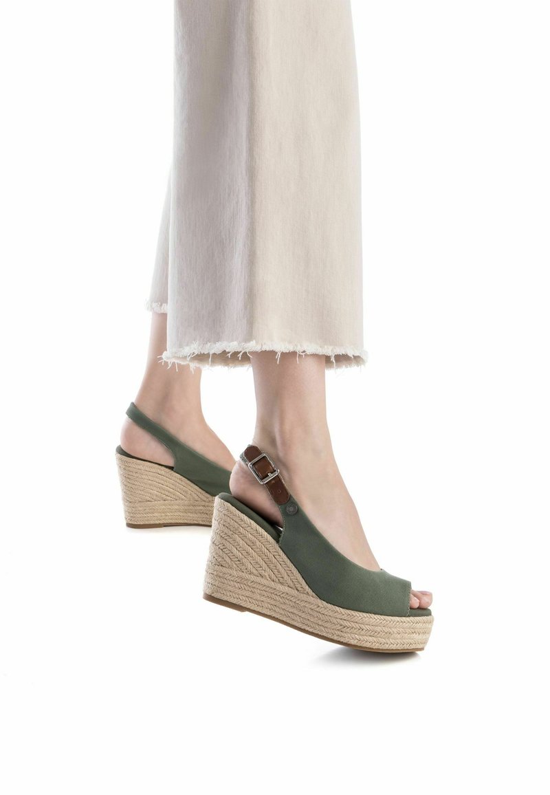 Sandalias verdes de cuña con diseño de punta descubierta, hechas de tela. Presentan un tacón de yute trenzado y una correa ajustable con hebilla. Se ven pantalones con dobladillo deshilachado.